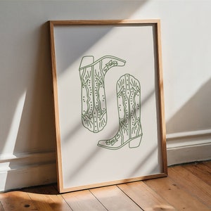 May include: A framed print showing a pair of green line-art cowboy boots. The boots have decorative stitching and are set against a neutral background. The artwork is displayed in a wooden frame.