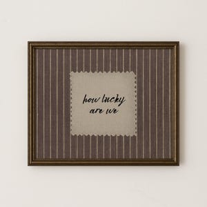 May include: Framed artwork featuring a brown and white striped pattern. A beige square with a decorative edge is at the centre, with the words "how lucky are we" in black script. The frame is a dark bronze colour.