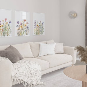 May include: A living room with a beige sofa, cushions, and a patterned throw. Three framed floral prints adorn the wall. A round wooden coffee table displays a vase with dried pampas grass. The room has a neutral colour scheme.