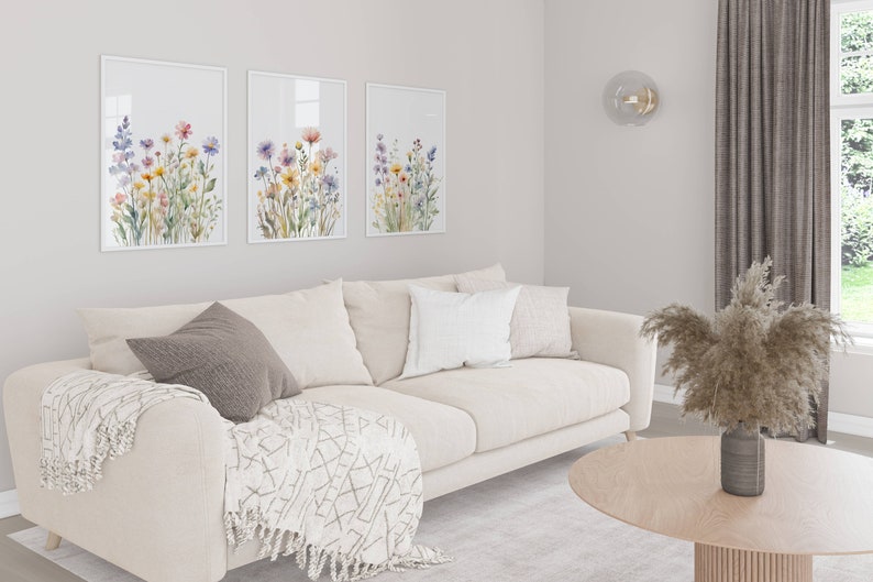May include: A living room with a beige sofa, cushions, and a patterned throw. Three framed floral prints adorn the wall. A round wooden coffee table displays a vase with dried pampas grass. The room has a neutral colour scheme.