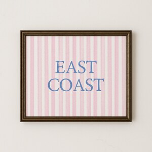 May include: Framed artwork with a brown border. The artwork features a pink and white striped background with the words "EAST COAST" in blue lettering. The artwork is rectangular and appears to be a print or poster.