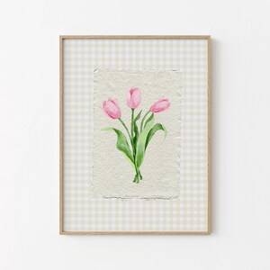 Pode incluir: Uma impress&atilde;o de arte emoldurada com uma ilustra&ccedil;&atilde;o em aguarela de tr&ecirc;s t&uacute;lipas cor-de-rosa com caules e folhas verdes sobre papel texturado branco sujo. A obra est&aacute; sobre um fundo de xadrez vichy bege claro e branco numa moldura de madeira.