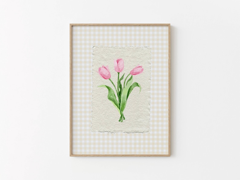 Pode incluir: Uma impress&atilde;o de arte emoldurada com uma ilustra&ccedil;&atilde;o em aguarela de tr&ecirc;s t&uacute;lipas cor-de-rosa com caules e folhas verdes sobre papel texturado branco sujo. A obra est&aacute; sobre um fundo de xadrez vichy bege claro e branco numa moldura de madeira.