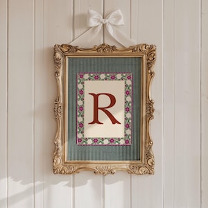 May include: A framed print featuring the letter "R" in a burgundy hue, surrounded by a floral border. The print is set against a cream background and a teal textured backdrop, encased in a gold ornate frame, and adorned with a white bow.