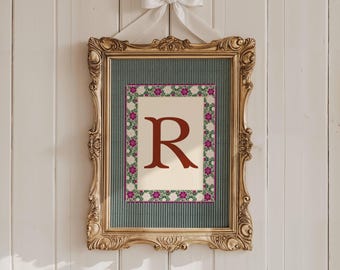 May include: A framed print featuring the letter "R" in a burgundy hue, surrounded by a floral border. The print is set against a cream background and a teal textured backdrop, encased in a gold ornate frame, and adorned with a white bow.