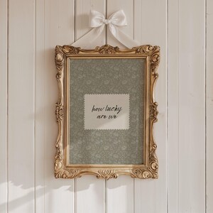 May include: Ornate gold picture frame with a floral patterned background and the text "how lucky are we." The frame is suspended by a white ribbon tied in a bow. The frame is hanging on a white wooden wall.