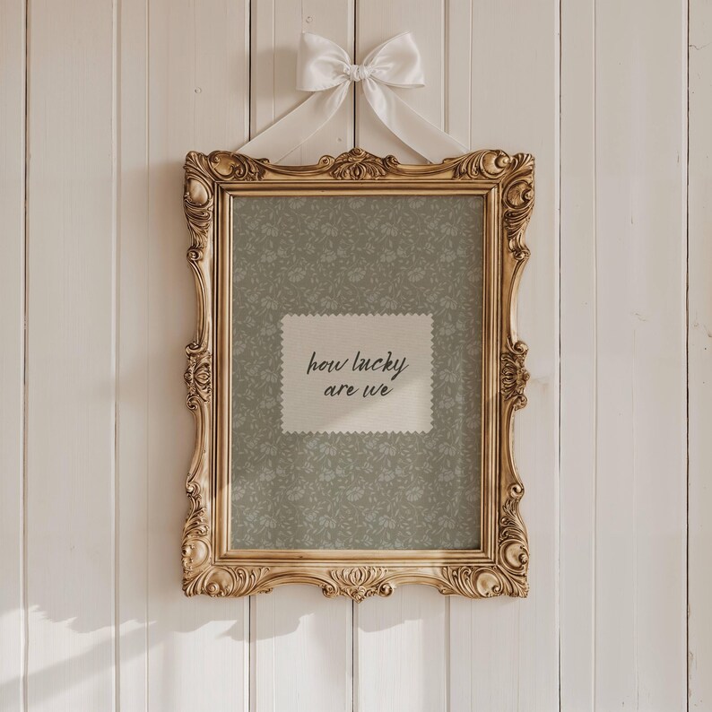 May include: Ornate gold picture frame with a floral patterned background and the text "how lucky are we." The frame is suspended by a white ribbon tied in a bow. The frame is hanging on a white wooden wall.