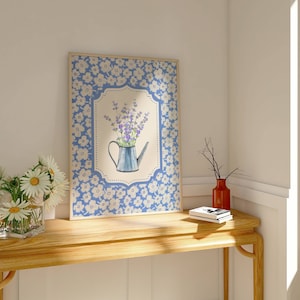 May include: A framed floral print featuring a blue watering can filled with purple flowers. The frame has a blue floral pattern. A vase of daisies and a red vase with branches sit on a wooden table.