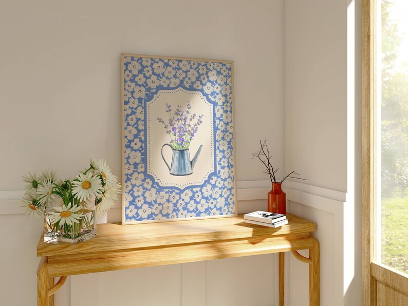 May include: A framed floral print featuring a blue watering can filled with purple flowers. The frame has a blue floral pattern. A vase of daisies and a red vase with branches sit on a wooden table.