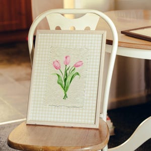 Pode incluir: Uma obra de arte emoldurada apresentando uma pintura em aquarela de tr&ecirc;s tulipas cor-de-rosa com caules e folhas verdes em papel texturizado branco sujo. A moldura tem uma borda clara com um padr&atilde;o xadrez vichy branco e verde claro.