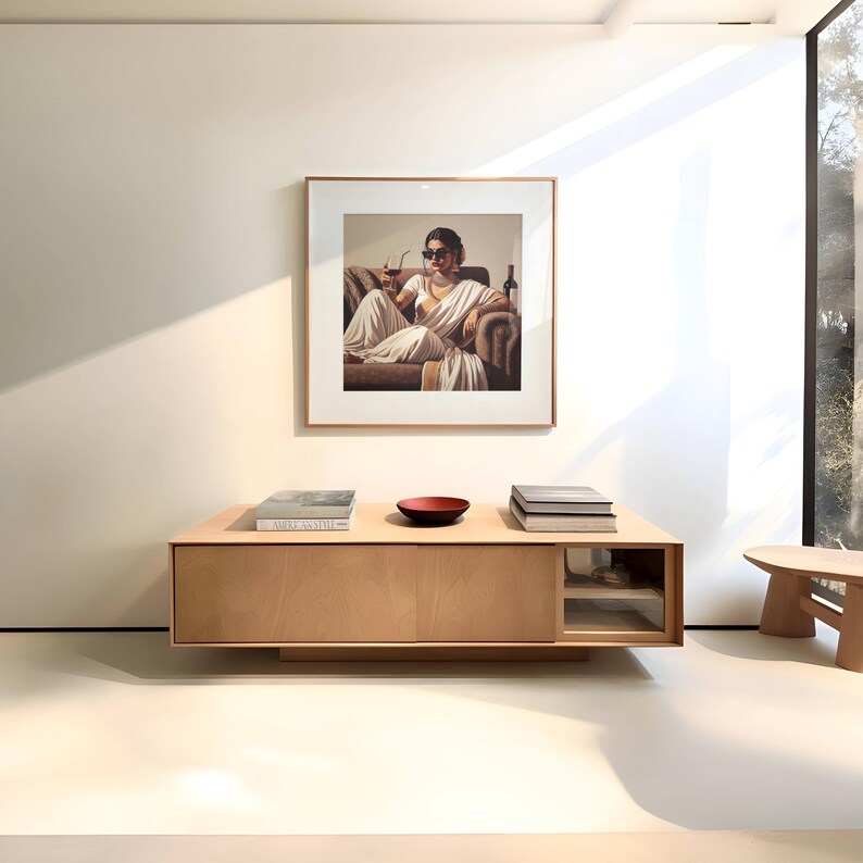 May include: A framed artwork depicts a person in a white sari, seated on a brown sofa, holding a wine glass. Below, a light wood cabinet displays books and a red bowl. The room has a minimalist design with natural light.
