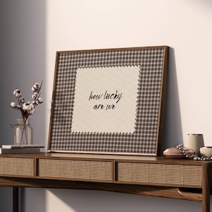 May include: A framed artwork with a brown and white checkered pattern border and the text "how lucky are we" on a cream-colored background. The artwork is displayed on a wooden console table with decorative items, including a vase of cotton stems.