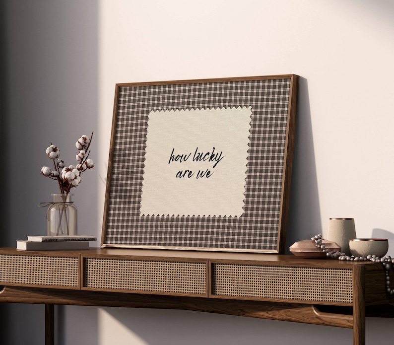 May include: A framed artwork with a brown and white checkered pattern border and the text "how lucky are we" on a cream-colored background. The artwork is displayed on a wooden console table with decorative items, including a vase of cotton stems.