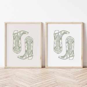 May include: Two framed prints each display a green line drawing of a cowboy boot. The boots are in a minimalist style with decorative stitching and the word "Evelyn" on the boot. One print has a cream background, the other a white background.