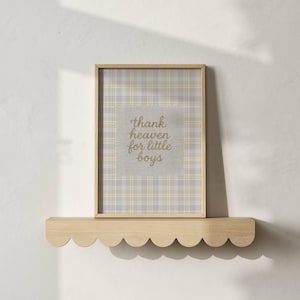 May include: A framed print with the text "thank heaven for little boys" in gold script. The print has a plaid pattern in neutral tones and is displayed on a scalloped wooden shelf.