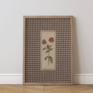 May include: A framed floral art print. The artwork features a botanical illustration of red flowers with green leaves. It is set against a brown and white plaid background and framed in natural wood. A decorative piece for home decor.