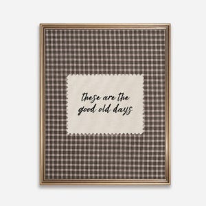May include: A framed print with a brown and white checkered pattern background. A cream-colored, scalloped-edge rectangle in the center features the handwritten text, "these are the good old days" in black script. The frame is gold.