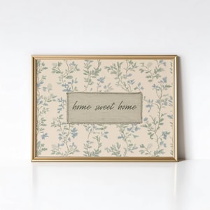 Op de afbeelding: Een ingelijste print met een goudkleurige lijst. De print heeft een bloemenpatroon in blauw en groen op een crèmekleurige achtergrond. Een rechthoekige patch in het midden leest "home sweet home" in een scriptlettertype.