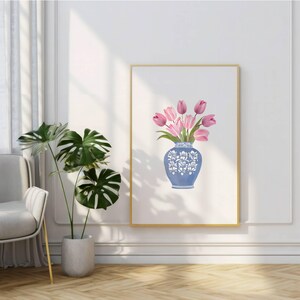 May include: A framed art print featuring a blue vase with white floral designs, holding pink tulips. The artwork is displayed on a white wall, with a gold frame. A potted plant with large green leaves is in the foreground.