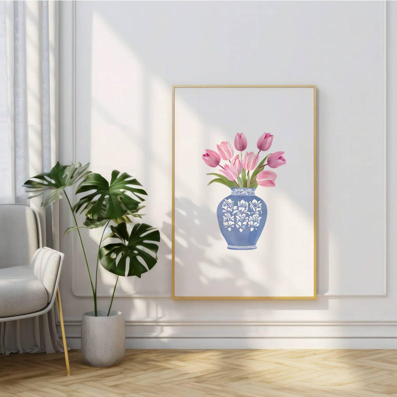 May include: A framed art print featuring a blue vase with white floral designs, holding pink tulips. The artwork is displayed on a white wall, with a gold frame. A potted plant with large green leaves is in the foreground.