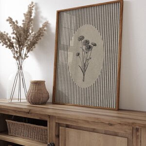 May include: A rectangular framed wall art piece featuring a vertical striped pattern in grey and white. Centered within the stripes is a beige oval containing a black line drawing of a cluster of wildflowers with thin stems and small petals.