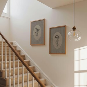 May include: Two rectangular wall art pieces in light wood frames, each featuring a vertical striped background with a central oval containing a black and white botanical illustration of flowers, displayed on a plain wall near a staircase.
