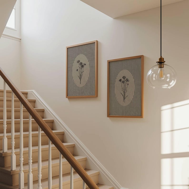 May include: Two rectangular wall art pieces in light wood frames, each featuring a vertical striped background with a central oval containing a black and white botanical illustration of flowers, displayed on a plain wall near a staircase.
