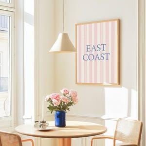 May include: A framed wall art print with pink and white vertical stripes and the words "EAST COAST" in blue. The print hangs above a round wooden table with a blue vase of pink flowers. A beige pendant lamp hangs above the table.