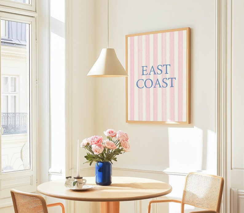 May include: A framed wall art print with pink and white vertical stripes and the words "EAST COAST" in blue. The print hangs above a round wooden table with a blue vase of pink flowers. A beige pendant lamp hangs above the table.