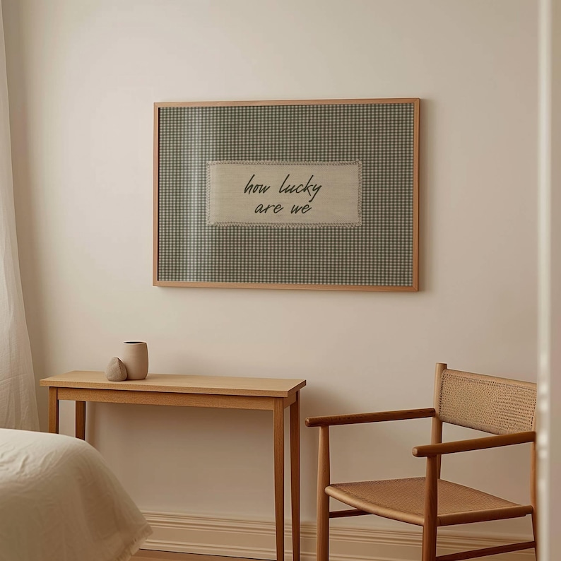 May include: A framed wall art piece with a gingham pattern background and a rectangular fabric patch in the center. The patch features the phrase "how lucky are we" in a handwritten style. The frame is made of light-colored wood.