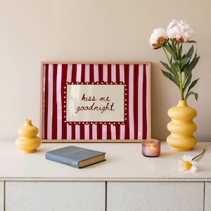 May include: A framed print with a red and pink striped background and the words "kiss me goodnight." The print is displayed on a white surface with a yellow vase holding flowers, a small candle, a book, and a small yellow vase.