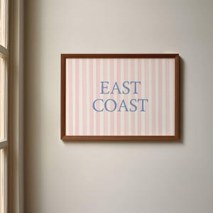 May include: A framed artwork with a brown wooden frame. The artwork features a pink and white striped background with the words "EAST COAST" in blue lettering. The artwork is hanging on a neutral-colored wall.