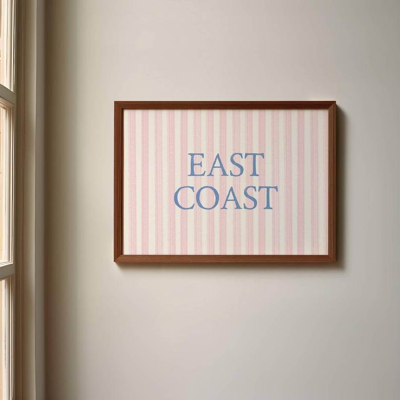 May include: A framed artwork with a brown wooden frame. The artwork features a pink and white striped background with the words "EAST COAST" in blue lettering. The artwork is hanging on a neutral-colored wall.