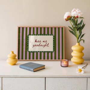 May include: A framed print with green and pink vertical stripes and the text "kiss me goodnight." The print is displayed with a yellow vase holding flowers, a small yellow vase, a blue book, and a lit candle in a glass holder.