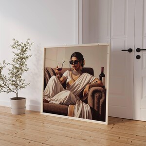 May include: A framed print depicts a woman in a white sari, sunglasses, and jewelry, seated on a brown sofa, holding a glass of red wine. A wine bottle sits nearby. The artwork is displayed against a white wall with a door and a potted plant.