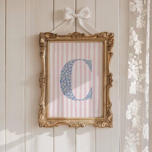 May include: A framed print featuring the letter "C" in a blue floral pattern, set against a pink and white striped background. The ornate gold frame is suspended by a white ribbon tied in a bow. The artwork hangs on a white wooden wall.