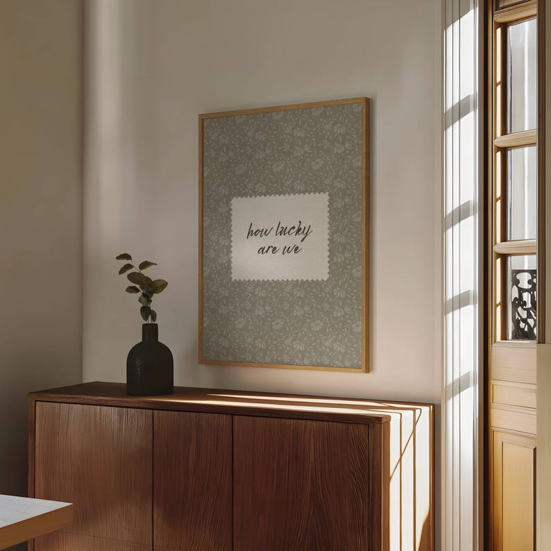 May include: A framed print with a floral pattern background and the text "how lucky are we" in a white square. The frame is a light wood color. A dark vase with greenery sits on a wooden cabinet.