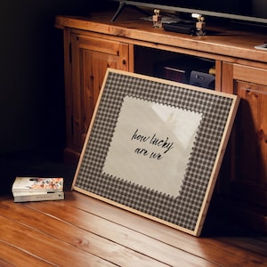 May include: A framed artwork with the text "how lucky are we" in a handwritten style. The frame has a brown and white checkered pattern. The artwork is leaning against a wooden cabinet.