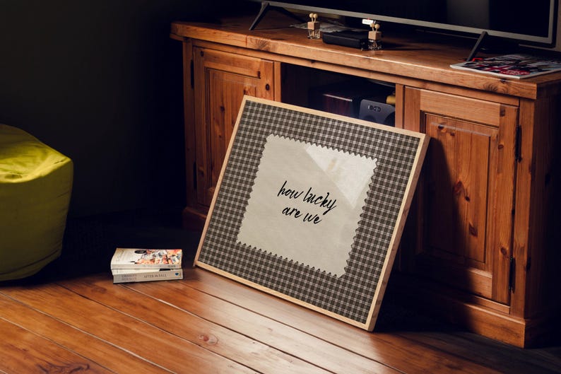 May include: A framed artwork with the text "how lucky are we" in a handwritten style. The frame has a brown and white checkered pattern. The artwork is leaning against a wooden cabinet.