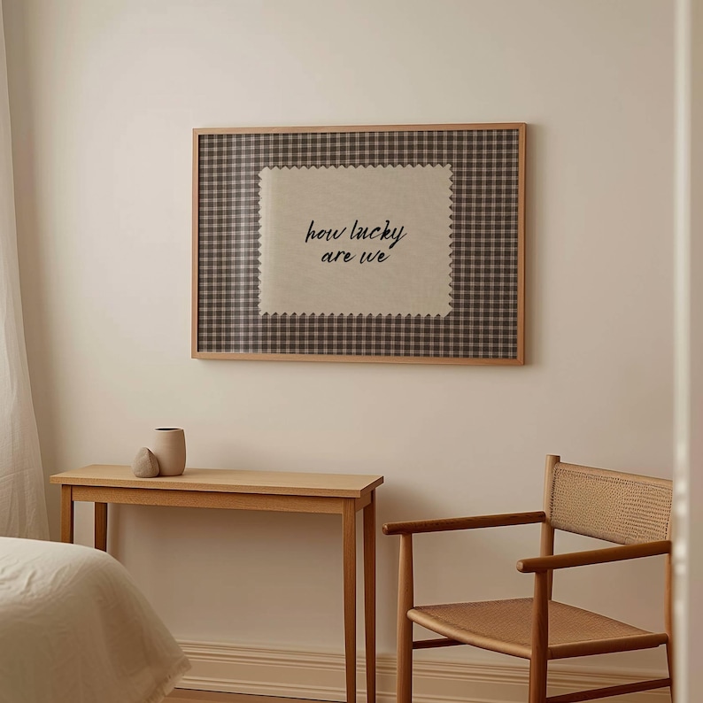 May include: A framed wall art piece with a gingham pattern and the text "how lucky are we." The artwork is displayed above a wooden table with a vase and a chair with a woven seat.