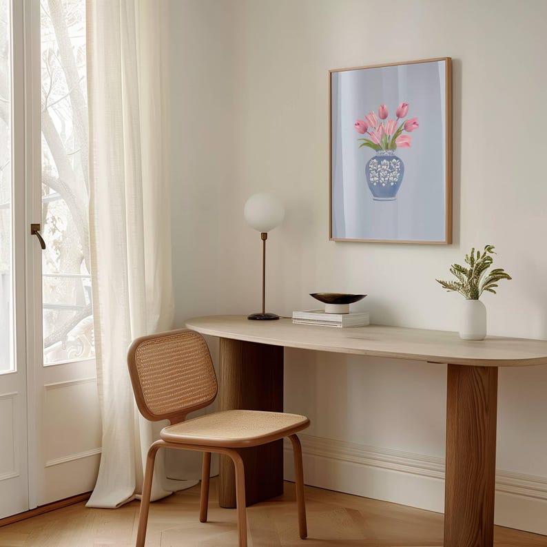 May include: A framed art print featuring a vase of pink tulips. The vase is blue with white floral designs. The print hangs above a light wood desk with a small lamp, a bowl, and a white vase with greenery. A woven chair is next to the desk.