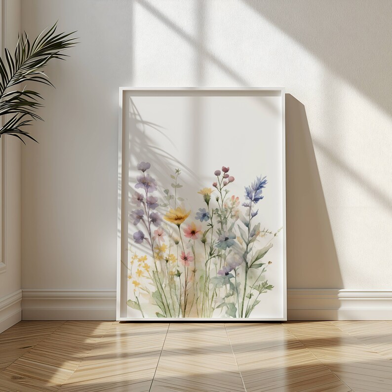 May include: A framed watercolour print depicting a colourful selection of wildflowers in shades of purple, yellow, pink, and blue. The artwork is set against a white background, with a white frame. The print is leaning against a white wall on a wooden floor.