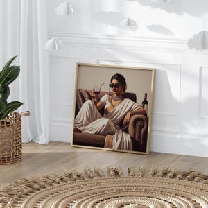 May include: Framed art print featuring a woman in a white sari, sunglasses, and jewelry, holding a glass of red wine. A bottle of wine sits beside her on a brown armchair. The artwork is in a gold frame, leaning against a white wall.