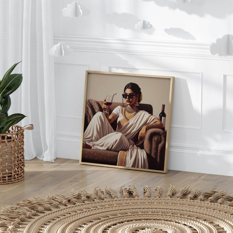 May include: Framed art print featuring a woman in a white sari, sunglasses, and jewelry, holding a glass of red wine. A bottle of wine sits beside her on a brown armchair. The artwork is in a gold frame, leaning against a white wall.