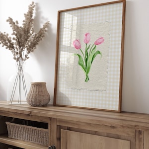 Pode incluir: Uma pe&ccedil;a de arte de parede emoldurada com uma ilustra&ccedil;&atilde;o em aguarela de tr&ecirc;s t&uacute;lipas cor-de-rosa com caules e folhas verdes em papel texturado. A impress&atilde;o est&aacute; sobre um fundo axadrezado bege claro e branco, numa moldura de madeira fina.