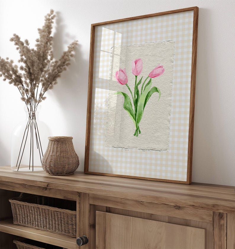Pode incluir: Uma pe&ccedil;a de arte de parede emoldurada com uma ilustra&ccedil;&atilde;o em aguarela de tr&ecirc;s t&uacute;lipas cor-de-rosa com caules e folhas verdes em papel texturado. A impress&atilde;o est&aacute; sobre um fundo axadrezado bege claro e branco, numa moldura de madeira fina.