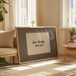 May include: A framed artwork featuring the phrase "how lucky are we" in black script. The artwork has a light wood frame and a gingham patterned background. The text is on a white, textured patch.