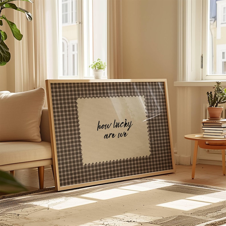 May include: A framed artwork featuring the phrase "how lucky are we" in black script. The artwork has a light wood frame and a gingham patterned background. The text is on a white, textured patch.