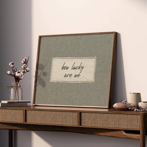 May include: A framed artwork with a green and white gingham pattern and the text "how lucky are we." The frame is made of dark wood. The artwork is displayed on a wooden console table with decorative items, including a vase of cotton stems.