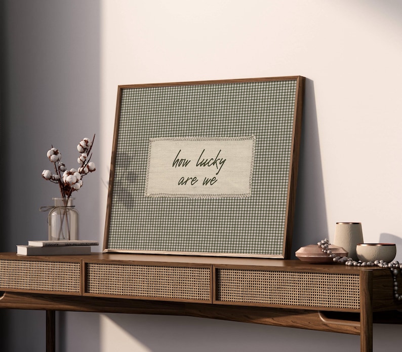 May include: A framed artwork with a green and white gingham pattern and the text "how lucky are we." The frame is made of dark wood. The artwork is displayed on a wooden console table with decorative items, including a vase of cotton stems.